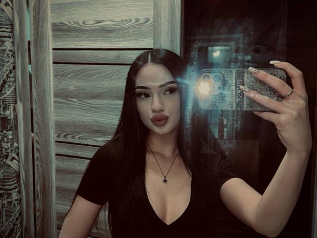 ShirleySiegel taking a selfie with a phone, showing her pouty lips, dark straight hair, black top, and a necklace, in a dimly lit wooden room. Boobs visible.