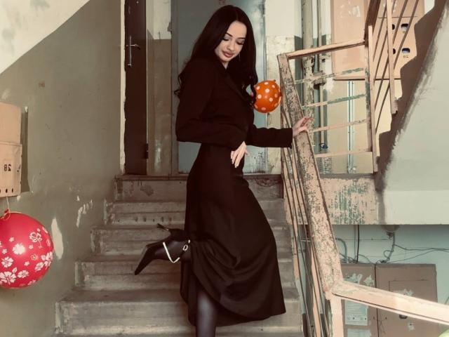ShirleySiegel with long black hair, black dress, and black boots, holding an orange balloon, standing on a concrete staircase, one boot lifted, showing her black panty.