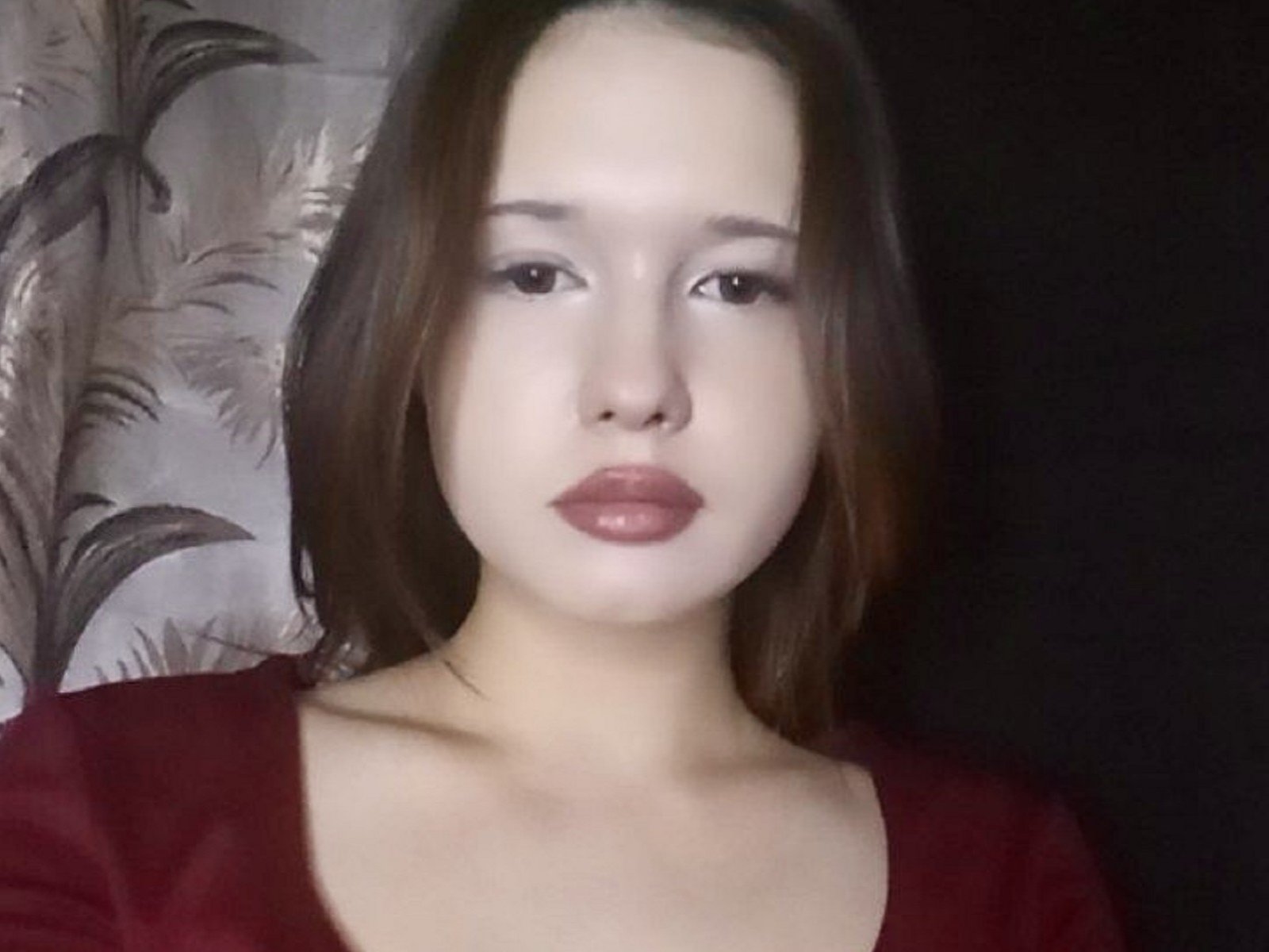 BambiBilinski with pale skin, brown eyes, and shiny pink lips, wearing a red top, has straight brown hair, looking slightly bored, in a soft light.