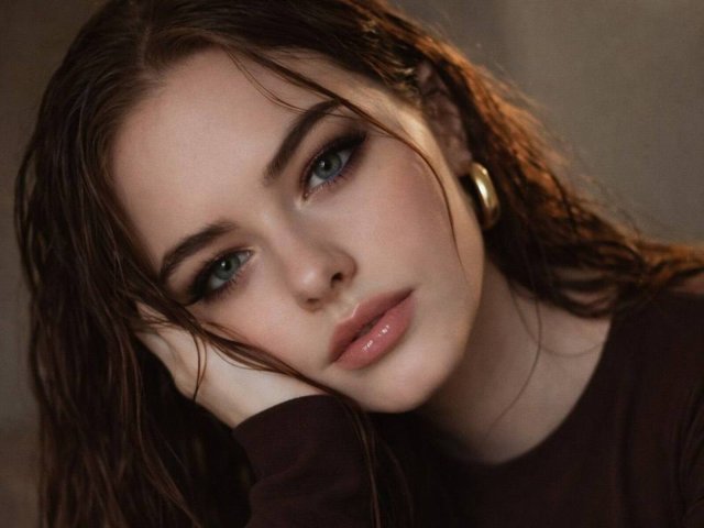 BryannaCrecelius with wet brown hair, green eyes, and glossy pink lips, resting her head on her hand, wearing gold earrings, in a dark top, looking pouty and sexy.