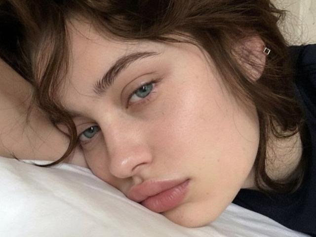 GrettaDemichelis with wavy brown hair, green eyes, and pink lips resting her head on a white pillow, looking slightly to the side with a calm expression.