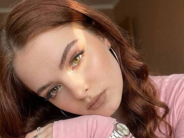 BryannaCrecelius has brown wavy hair, green eyes, and full pink lips, wearing a pink shirt, with a silver watch and black earbuds. She's looking at the camera.