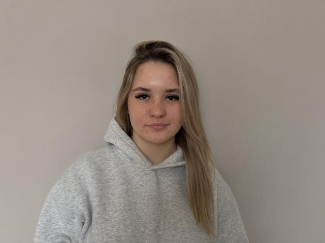 IzolaKlinglesmith has blonde hair, light freckles, and is wearing a gray hoodie, looking straight at the camera with a slight, cute smile. Her tits are small and subtle.
