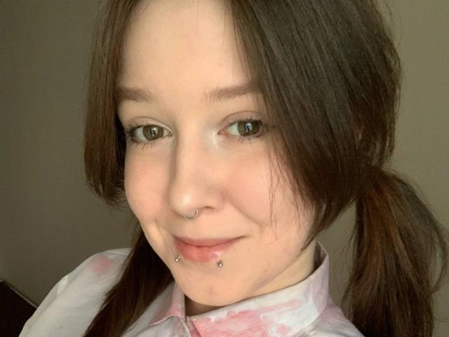 RubyOiler with brown pigtails, nose ring, and two lower lip piercings, smiling at the camera, wearing a pink and white tie-dye shirt.
