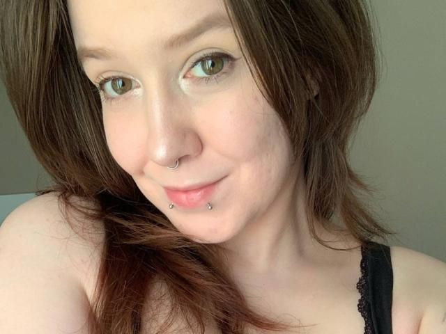 RubyOiler has brown eyes, a nose ring, and a lip piercing with small studs. She's smiling with light skin and brown hair, wearing a black lace top.