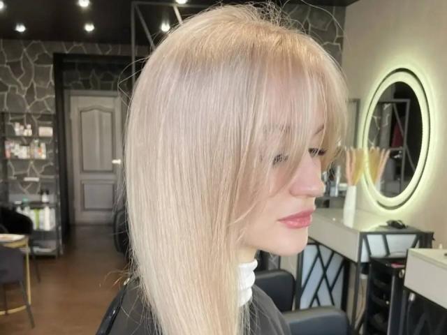 Margery Doster a de longs cheveux blonds et lisses avec une frange, une peau claire et du rouge à lèvres rose. Elle est assise dans un salon de coiffure, avec un miroir et un fauteuil noir en arrière-plan..
