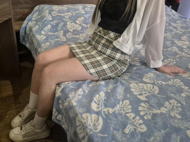 ZoieHurt sitting on a bed, wearing a plaid skirt, white shirt, and sneakers, with her legs spread and hands on the bed, showing her bare thighs and butt.
