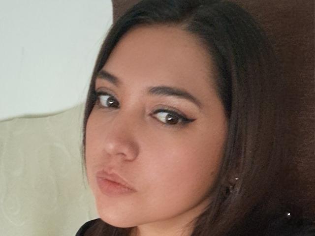 PaulaAnd has brown eyes, full lips, and straight dark brown hair, wearing black eyeliner, with a subtle, pouty expression, and small earrings on her left ear.