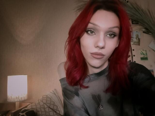 KiranaHayes with red hair, green eyes, and full lips, wearing a black tie-dye shirt, and a necklace, has a neutral expression, sitting in a softly lit room.