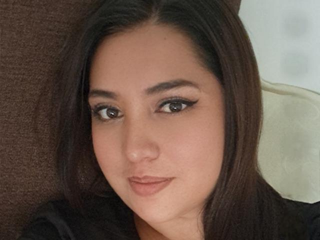 PaulaAnd has brown eyes, black eyeliner, and straight brown hair. She's smiling with a slight pout, wearing a black top. Her ass is not visible.