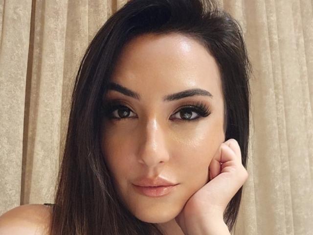 AlexaTailor hat glänzende braune Augen, dicke schwarze Wimpern und einen leichten Schmollmund, wobei ihre Hand unter ihrem Kinn liegt. Sie blickt direkt in die Kamera mit glatter, strahlender Haut..