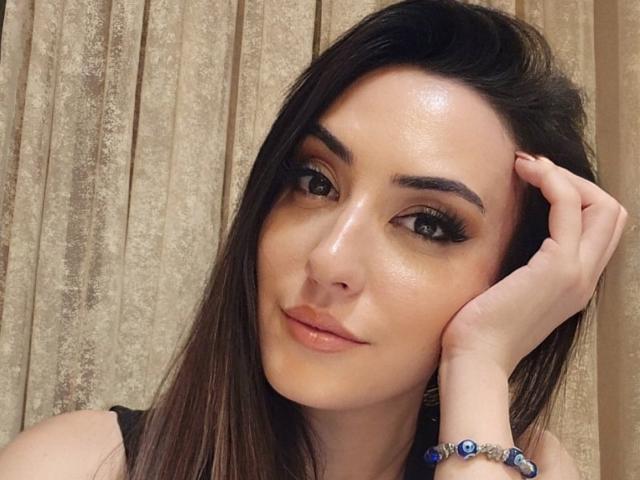 AlexaTailor with shiny skin, dark eyebrows, long eyelashes, and brown hair, touching her head with a hand, wearing a blue bracelet, smiling slightly.