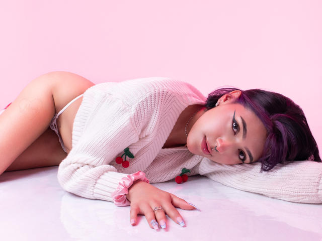 DulceKane is lying on her side with her ass up, wearing a white knitted sweater, pink scrunchie, and white thong, with cherry decorations. She has purple hair and a nose ring.