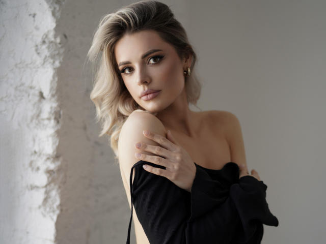 MoonShineX with blonde hair, holding black shirt over bare tits, covering ass, looking at camera, soft light, subtle makeup, gold earrings, textured white wall in background.