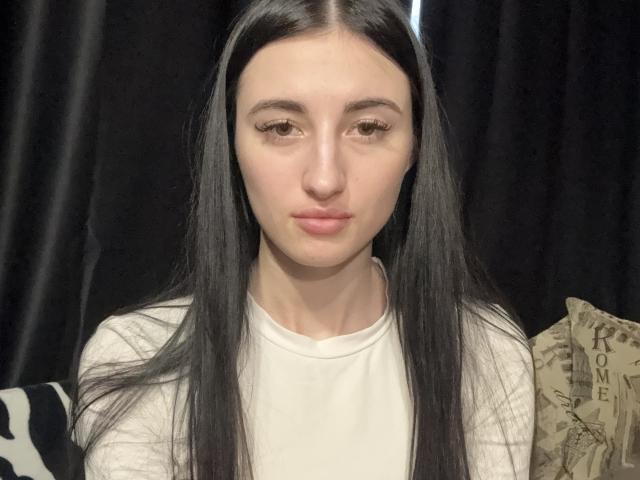 LanetteTaro with long black hair, pale skin, and brown eyes, wearing a white shirt, has a neutral expression and is looking slightly to the side in a selfie.