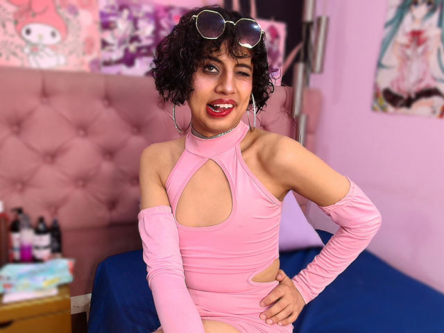 EimyFlorian with curly black hair, wearing a pink cutout dress, pink arm sleeves, and sunglasses on her head, smiling with red lipstick, showing her small tits and toned arms.