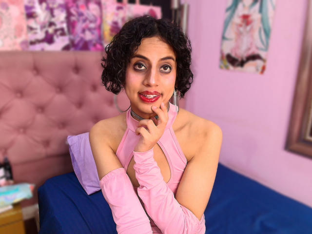 EimyFlorian with curly black hair, pink dress, and choker, touching her chin with a finger, blue eyes, red lipstick, and a cute smile, sitting on a blue bed.