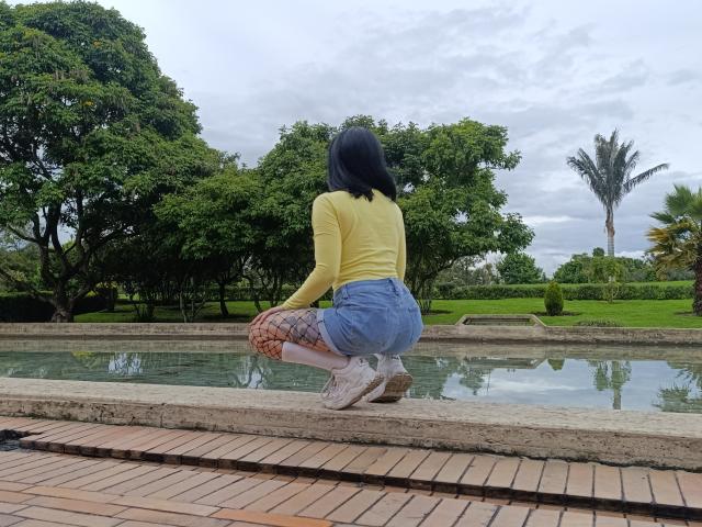 CandyParis crouches by a water feature, wearing a yellow long-sleeve top, blue shorts, snake-pattern leggings, and white sneakers, with her black bobbed hair and butt facing the camera.