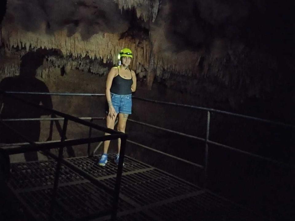 MarJoana, avec un casque jaune, un débardeur noir, un short bleu et des chaussures bleues, debout sur une grille métallique dans une grotte sombre, une ombre sur le mur, tenant une rambarde, regardant vers l'avant..