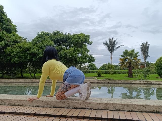 CandyParis on all fours, yellow top, blue shorts, fishnet stockings, white sneakers, black hair, yellow skin, over a brick-lined pool, with green trees and palm trees in the background.