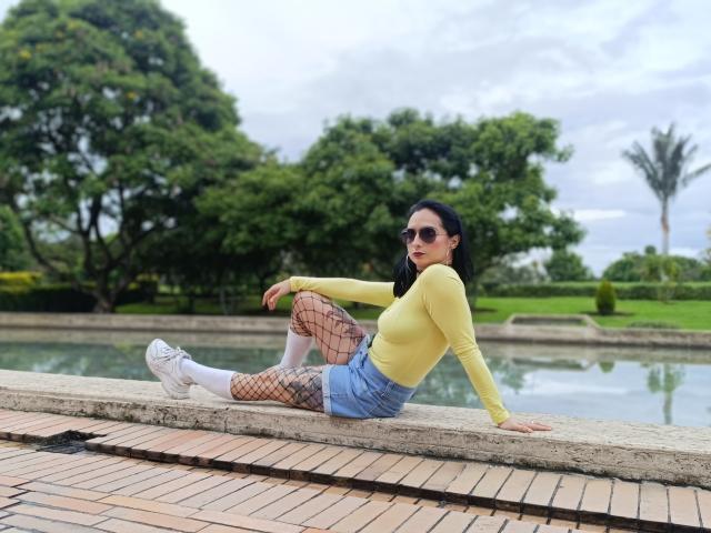 CandyParis sitting on a brick ledge, wearing a yellow long-sleeve top, blue shorts, fishnet tights, white socks, and white sneakers, with black pigtails and sunglasses.