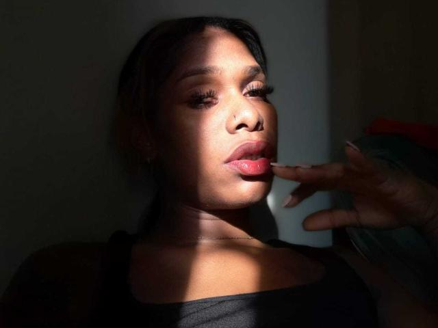 Reine guerrière à la peau foncée, aux lèvres roses et brillantes, et aux cheveux noirs, la lumière du soleil projetant une ombre sur son visage, un doigt effleurant délicatement sa lèvre, portant un haut noir, affichant une confiance et une sensualité..