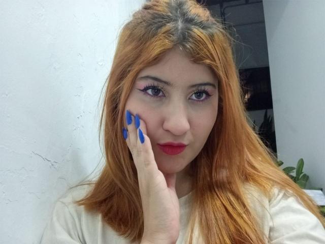 Beste Selene, met oranje haar, blauwe nagels, roze eyeliner en rode lippenstift, die haar wang aanraakt met een hand, een witte blouse draagt en met een lichte glimlach naar de camera kijkt..