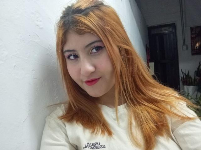 DearSelene has light brown hair, red lipstick, and a cute smile. She's wearing a white shirt with a text logo. Her eyes are slightly upturned, and she's looking at the camera.
