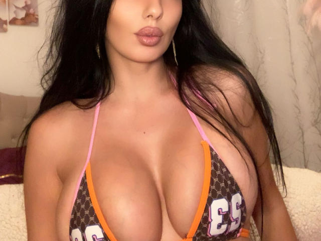 BrunetteBabe69 with large tits in a brown and orange sports bra, pink straps, long black hair, pouty lips, and gold hoop earrings. Bedroom setting, soft lighting.