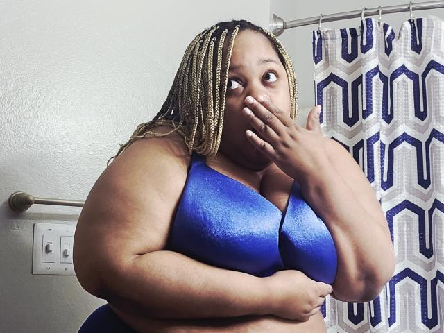 MorganMinx, de talla grande, con cabello rubio y marrón trenzado, cubriéndose la boca en un baño, vistiendo un sujetador azul brillante, con una expresión de sorpresa. Sus grandes senos son visibles..