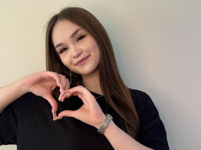 HettieBogdanovich, with long brown hair, black shirt, and silver watch, makes a heart shape with her hands, smiling gently at the camera.