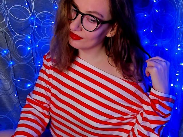 PaulinaSunny with red and white striped shirt, black glasses, red lipstick, and brown wavy hair, holding hair with right hand, blue fairy lights in background.