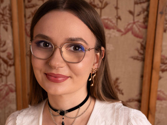 EllyBrune has glasses, blue eye makeup, and a black choker with chains. She's wearing a white top, gold earrings, and has a cute smile. Her tits are small.
