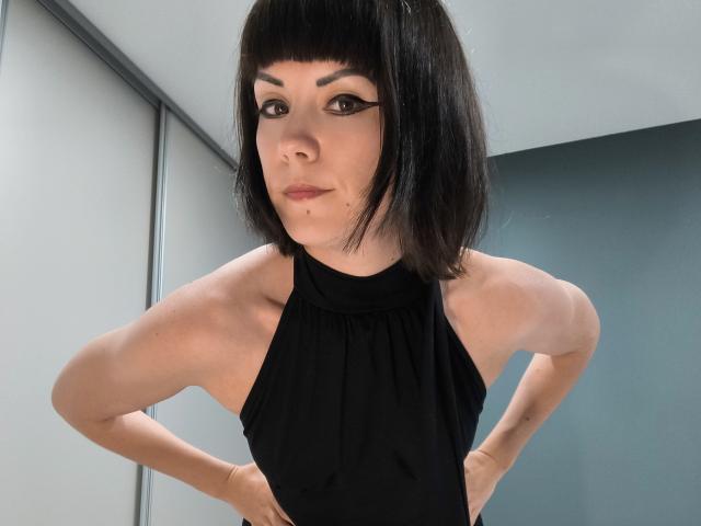 OrianaLaFrancaise with black bob, eyeliner, and black halter top, arms folded, showing muscular arms and small tits, standing in a room with gray and white walls.