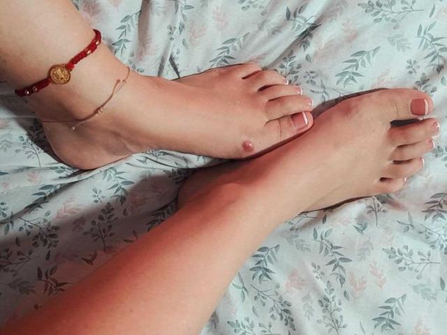 Kiaa69's bare feet with pink toenails, red beaded anklet, and gold bracelet, ankles crossed on floral bedsheet. Her small, pink nipples are visible between toes.