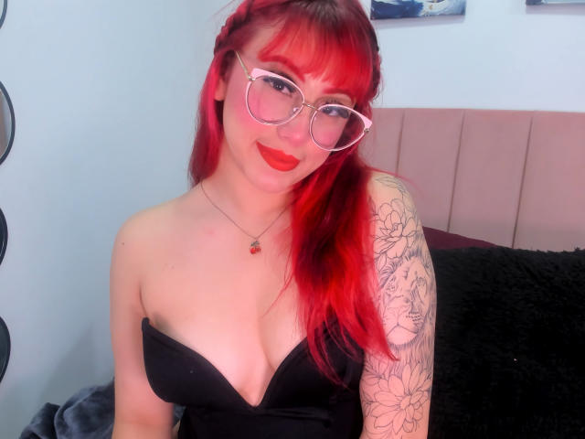 MaddyMills with red hair, glasses, and red lipstick, showing off her tattoos and black strapless dress, showcasing her boobs. She's smiling with a cute, sexy look.