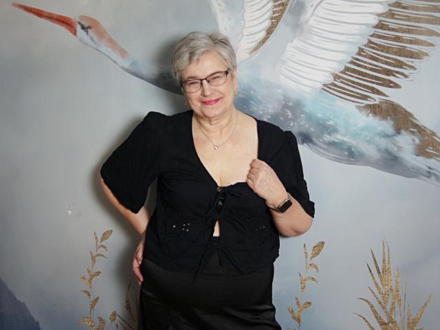 SensualIris, elderly woman with short gray hair, glasses, black button-up top, black skirt, pink lipstick, smiling, right hand on hip, left hand on chest, wearing a wristwatch.