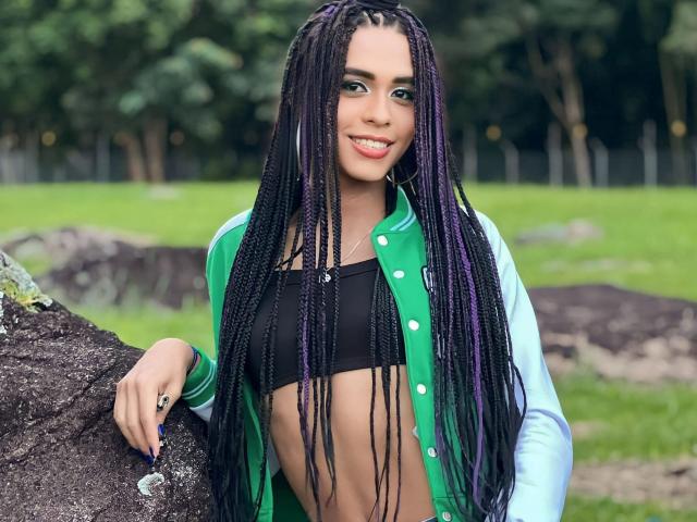 SexyHannaD with long purple and black braids, wearing a green and blue jacket over a black crop top, leaning on a rock, smiling with cute, green eyes.