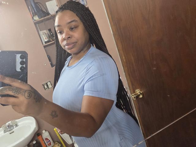 RosenBudd with dark skin, long braids, wearing a light blue ribbed shirt, holding a phone, showing tattoos on her right hand, standing in a bathroom with a wooden door.