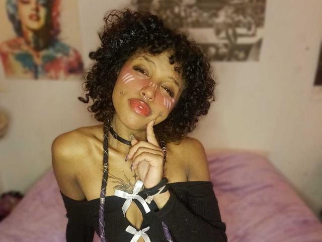 Luna Keller, com cabelos cacheados, maquiagem branca no rosto, piercing no nariz e blusa preta com um ombro de fora e fita branca, fazendo um gesto com os dedos, mostrando seus lábios grossos e seios pequenos..