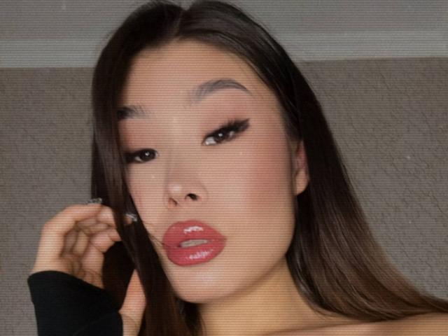 AizaXo's face with long brown hair, red glossy lips, and black eyeliner, showing off her shiny ass, with a black sleeve and her hand near her face.