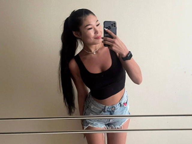 AizaXo with long black ponytail, black tank top, and denim shorts, holding a phone, smiling, wearing a choker and wristwatch, standing in front of a mirror.