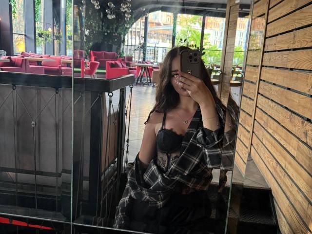 Mikkinik met lang bruin haar, een zwarte kanten bh en een geruite blouse, maakt een spiegelzelfportret, waarbij haar borsten en telefoon zichtbaar zijn in een houten restaurant met rode tafels..