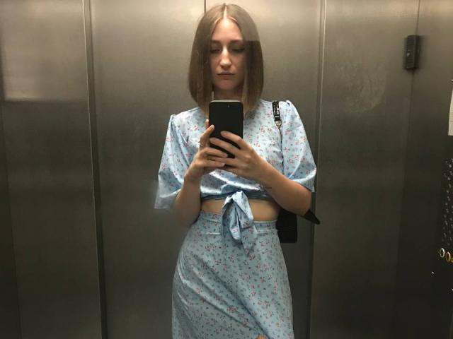 EvvaSmit, dans un autoportrait pris dans un miroir, porte une robe fleurie bleu clair nouée, qui révèle ses petites seins et ses fesses plates, et tient un téléphone noir dans un ascenseur métallique..