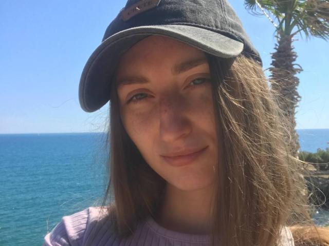 EvvaSmit with long brown hair, wearing a grey cap, smiling at the camera, blue ocean in the background, wearing a pink ribbed shirt, sunny day, palm tree on right.