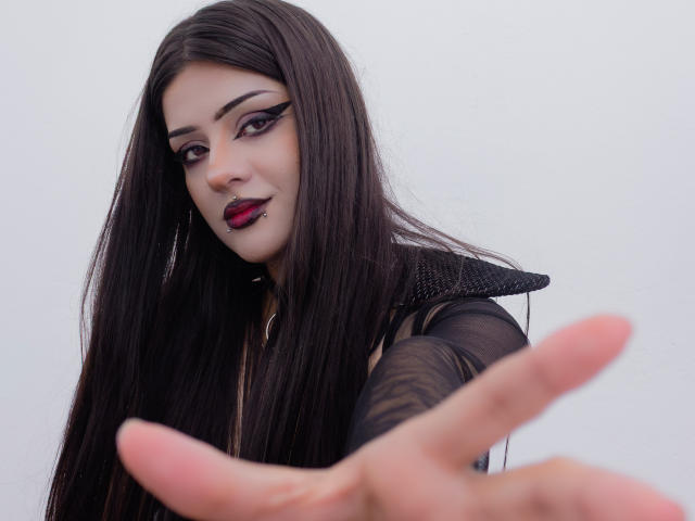 HelenaMorningstar with long black hair, goth makeup, red lipstick, black eyeliner, black dress, reaching out with her right hand, pale skin, dark eyebrows, looking at the camera.