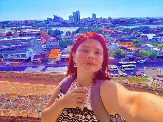 KatherineToro has red hair, wearing a black-and-white patterned dress, smiling with a hand near her chest, holding a backpack, and looking at the camera from a rooftop.