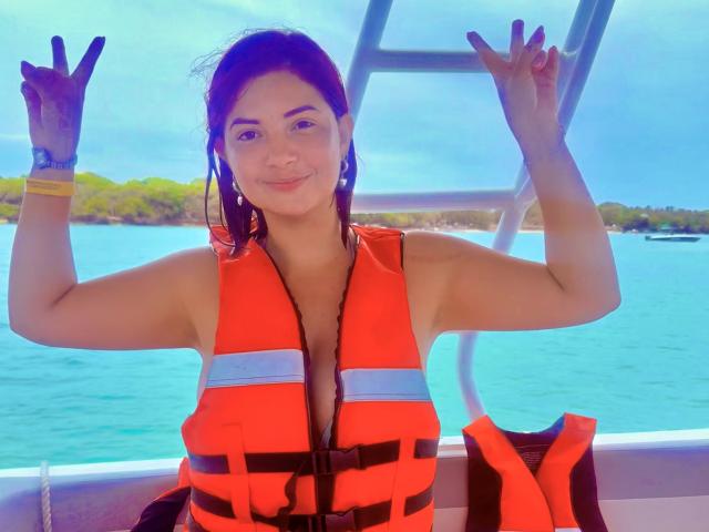 Katherine Toro, com seios médios, vestindo um colete salva-vidas laranja, fazendo dois sinais de "paz" em um barco, sorrindo, com cabelo escuro, usando uma pulseira amarela, com um fundo de água azul..