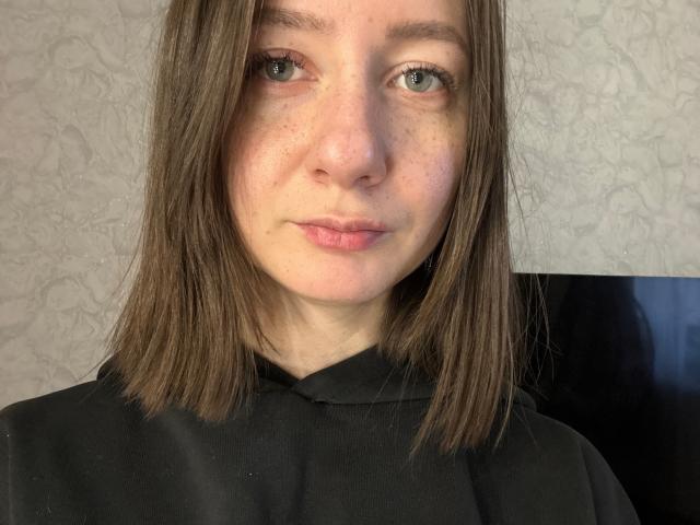 EnidBartuska has medium-length brown hair, green eyes, freckles, and a subtle smile. She's wearing a black shirt, looking straight at the camera with a neutral expression.