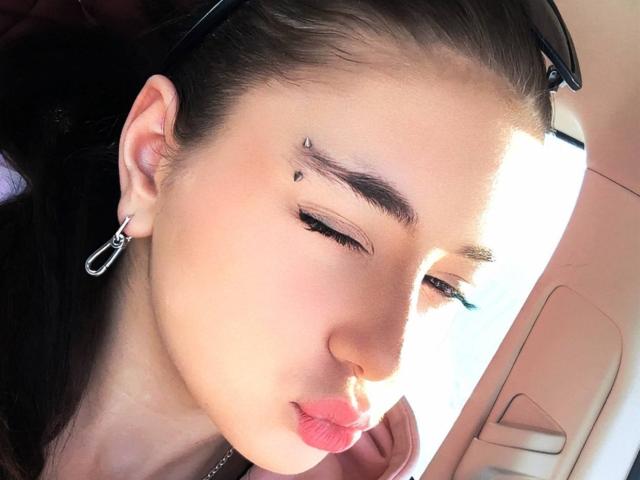 Le visage de JaninaVidrio, peau brun clair, cheveux brun foncé, eye-liner noir, petite tache au-dessus du sourcil droit, lèvres roses, boucles d'oreilles noires, lunettes de soleil noires sur la tête, intérieur de voiture, lumière du soleil..